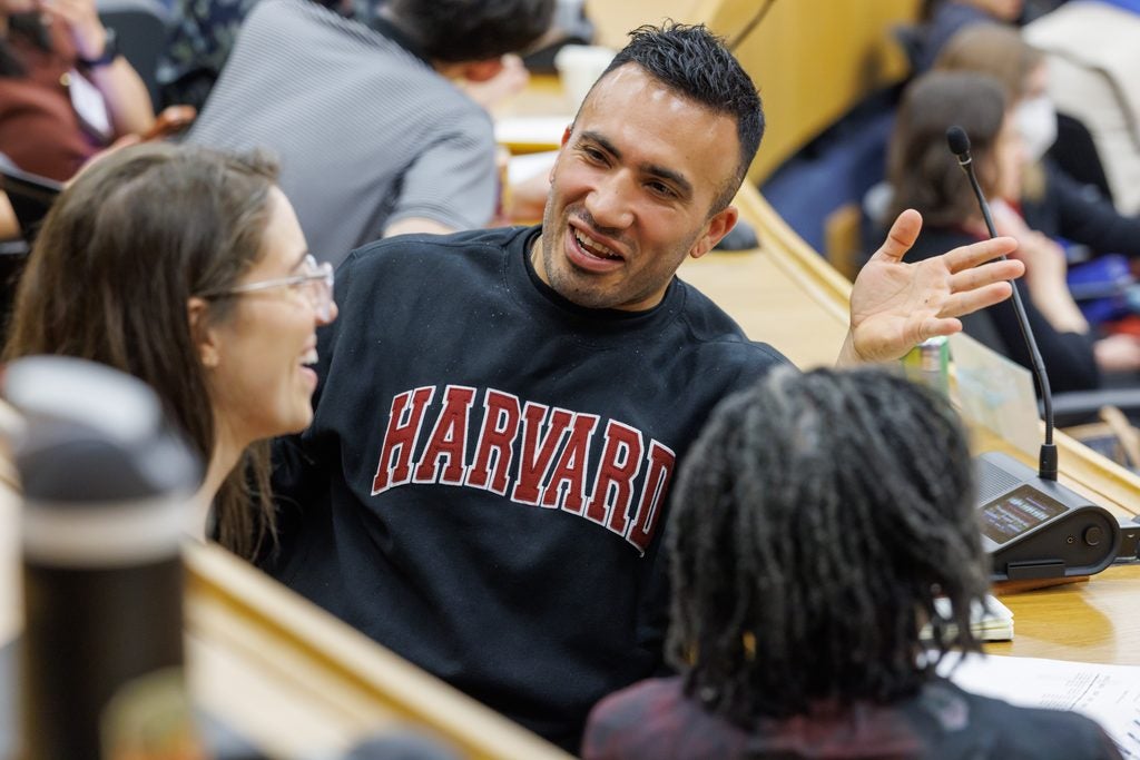 A man in a Harvard sweatshirt is turned, speaking animatedly to his seat neighbors.