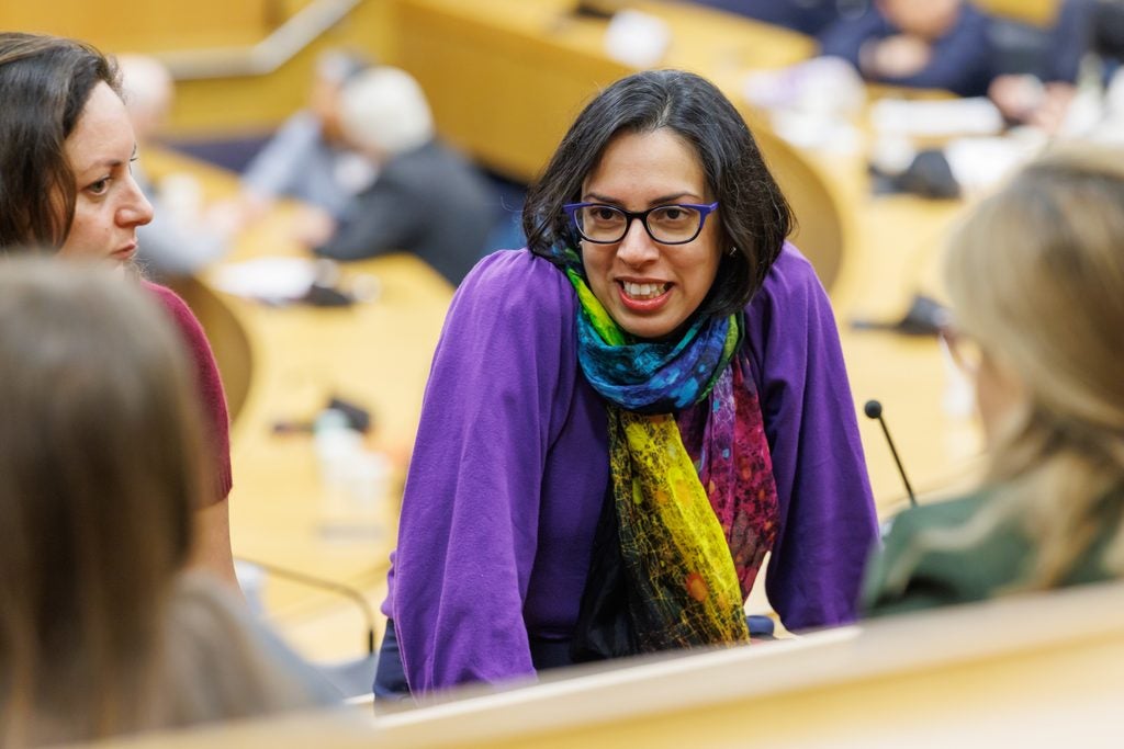 A woman listens in the lecture hall, wearing a striking purple blouse, rainbow scarf, an purple-framed glasses. 