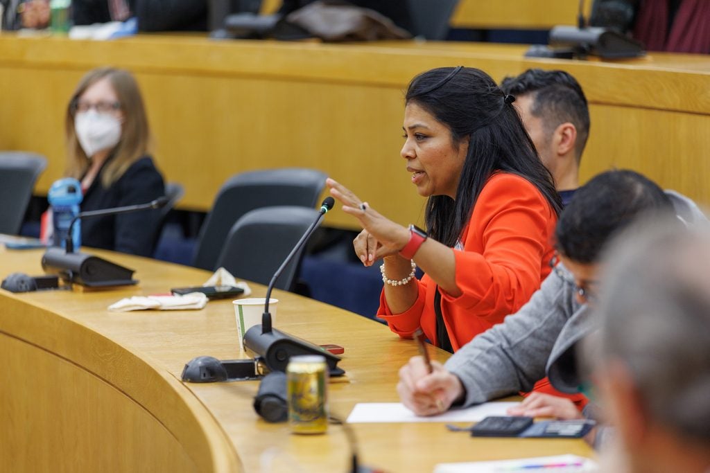 A woman in a bright orange blazer speaks from her lecture hall seat., 