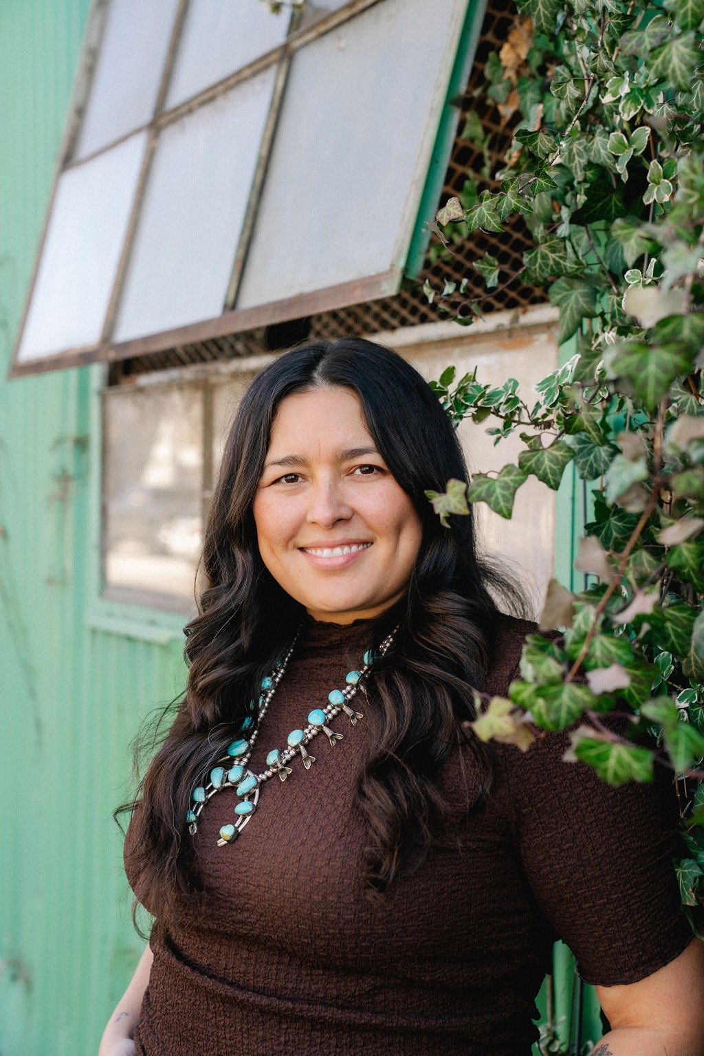 Brit Koch in a dark purple shirt from the waist up. A green wall with a window is behind her.