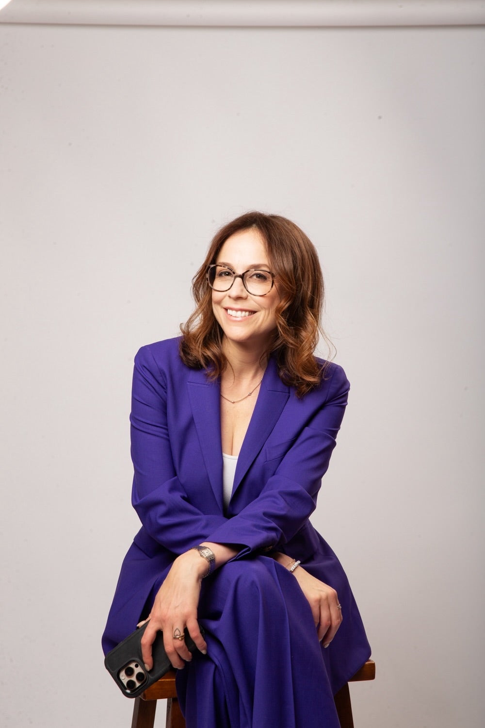 Shira Lazar wearing a purple suit and glasses against a warm grey backdrop.