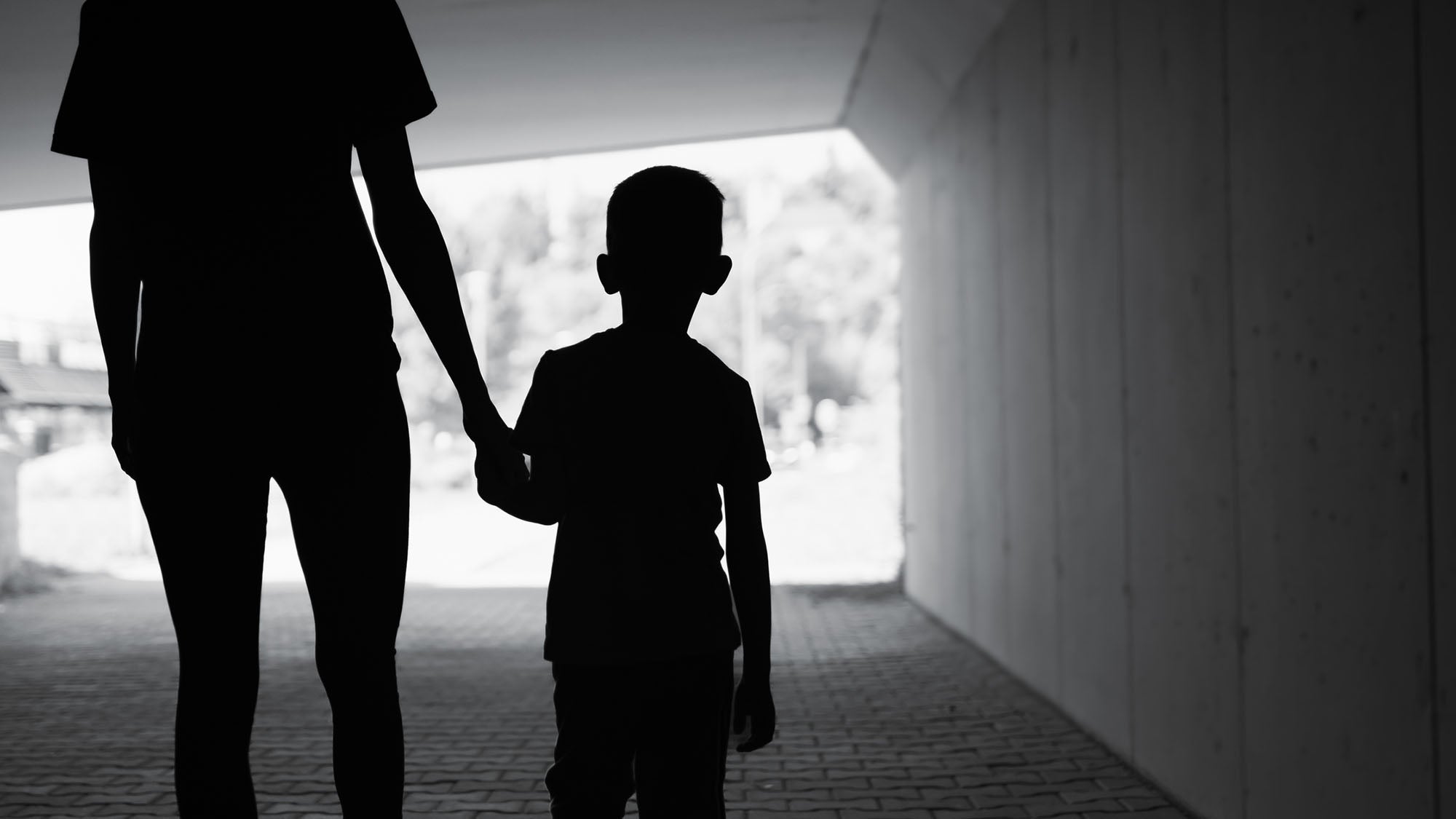 Silhouette of mother holding son's hand, walking on a city street, under an overpass.