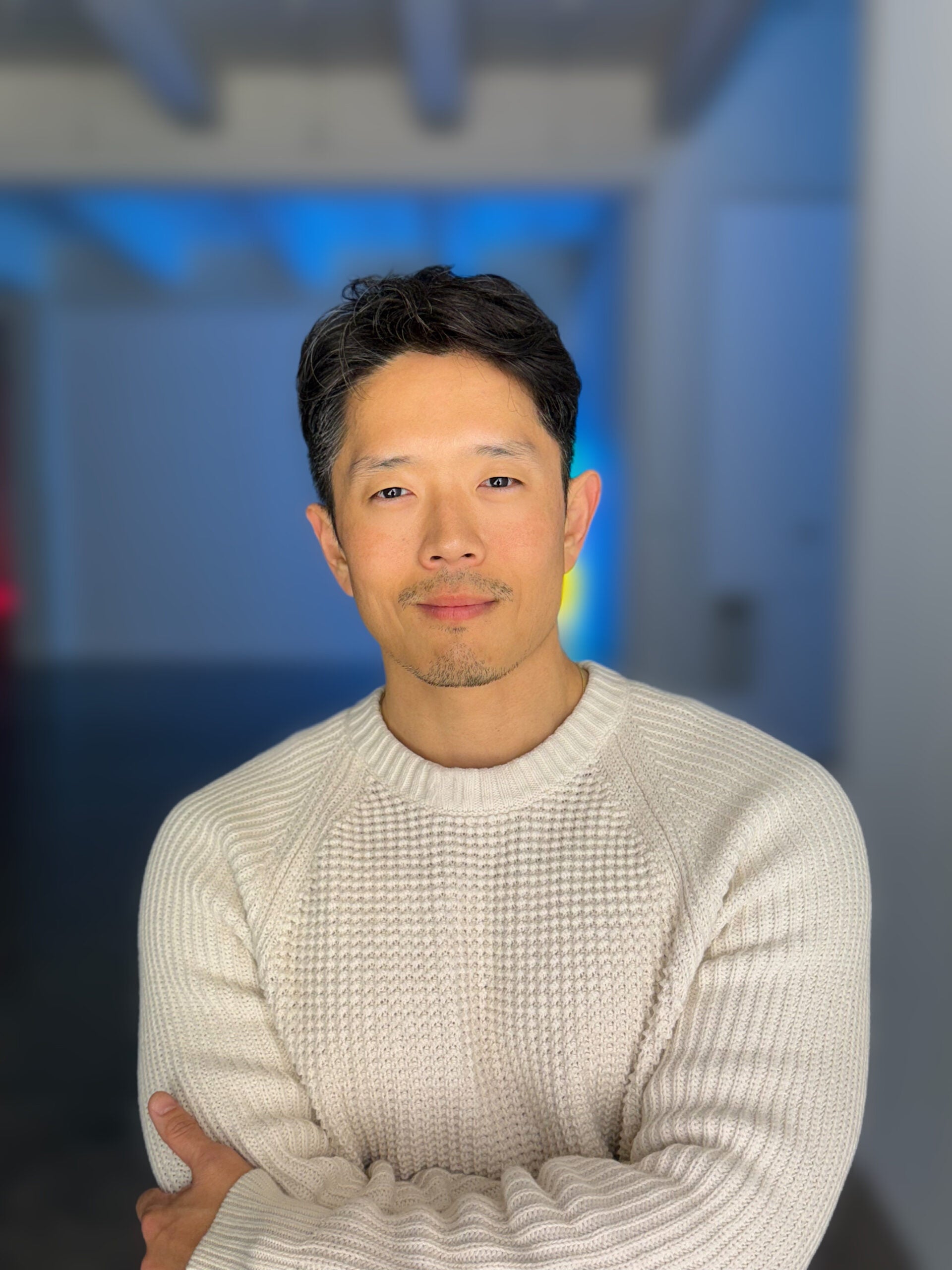 Nathan Jun in a white sweater with a empty room with blue lighting behind him.
