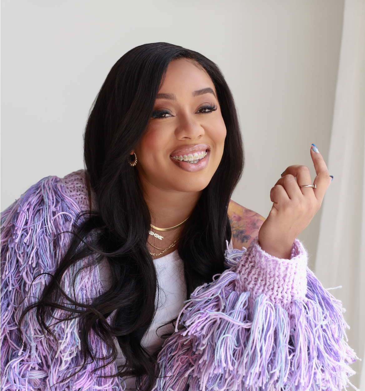 Jouelzy is wearing a fuzzy purple sweater and gold necklaces against a beige wall.