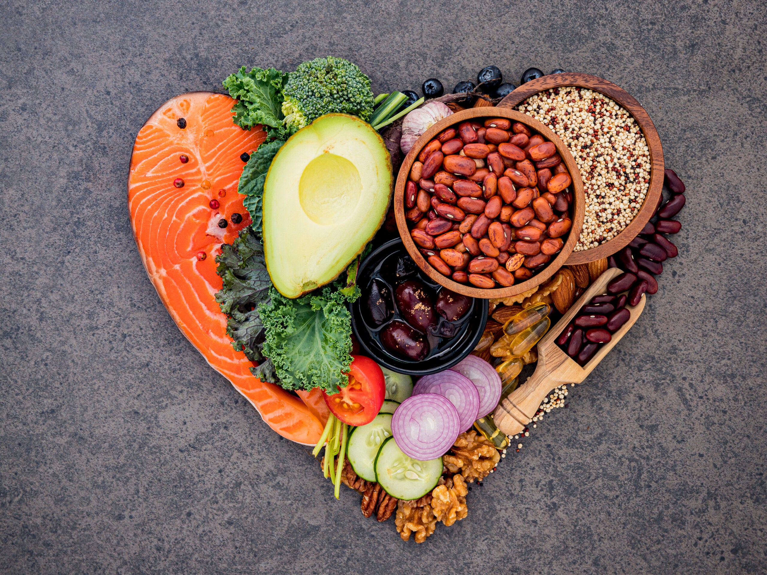 Healthy foods, such as salmon, avocado, kale, nuts, beans, and blueberries, arranged together to form the shape of a heart.