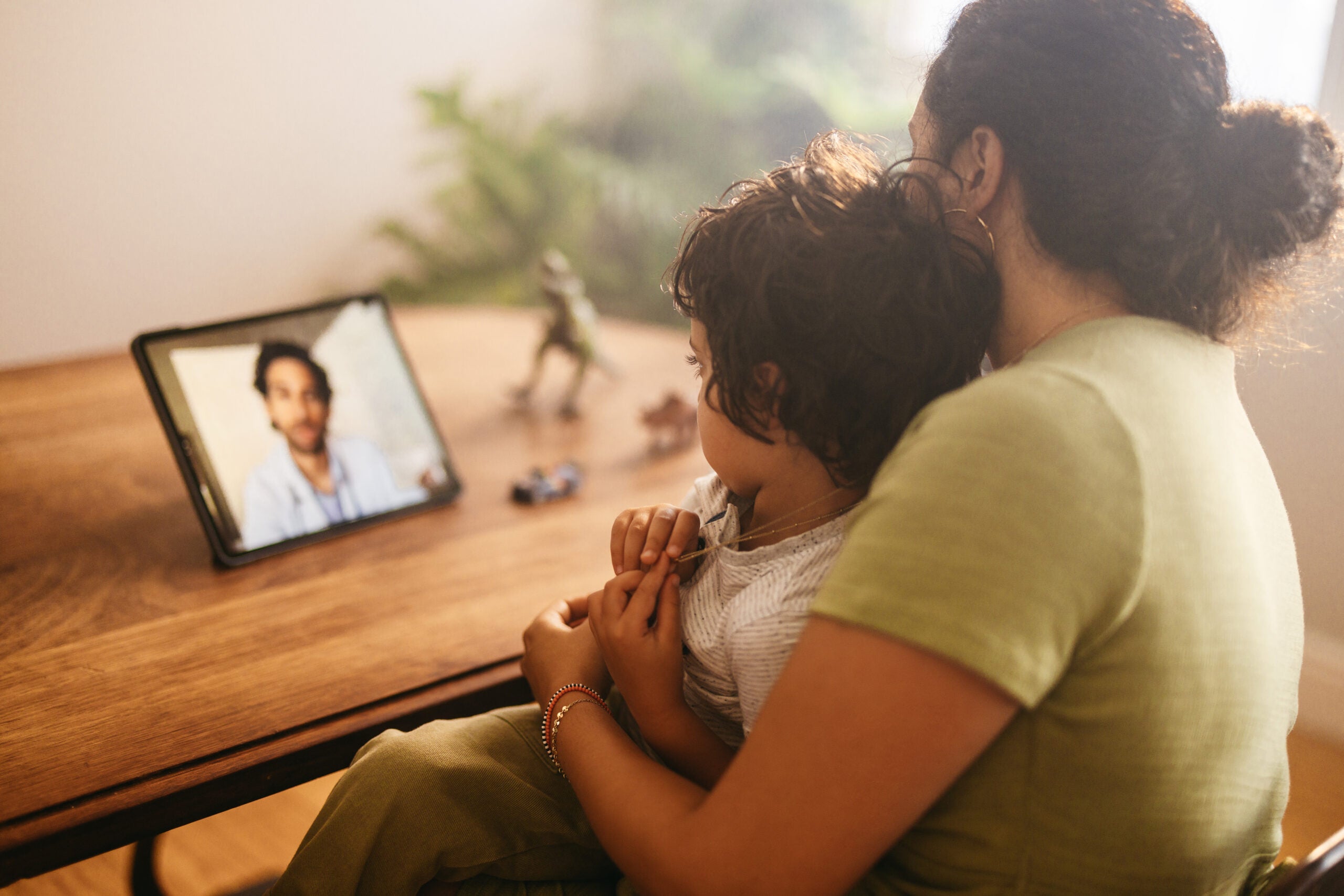 A boy sits on his mother's lap during a telehealth visit. They're photographed from behind. The clinician is pictured through a tablet device on the table.