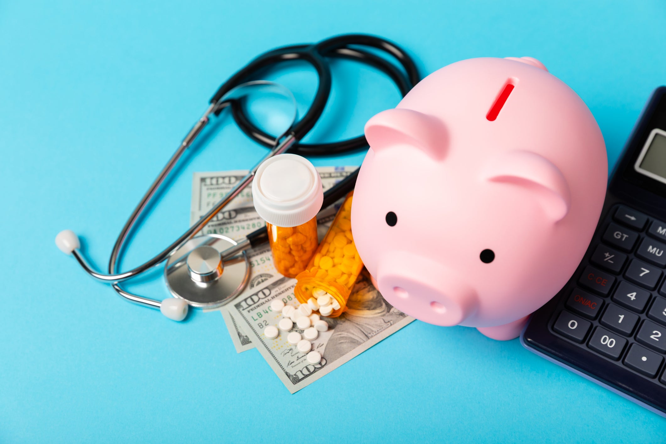 Still life photo of a piggy bank surrounded by dollar bills, a stethoscope, pill bottles, and a calculator.